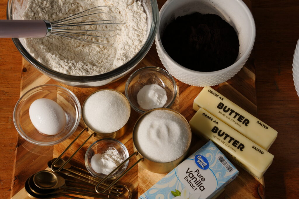 Overhead view of cookie ingredients including flour, cocoa powder, butter, sugar, egg, and vanilla on a wooden board.