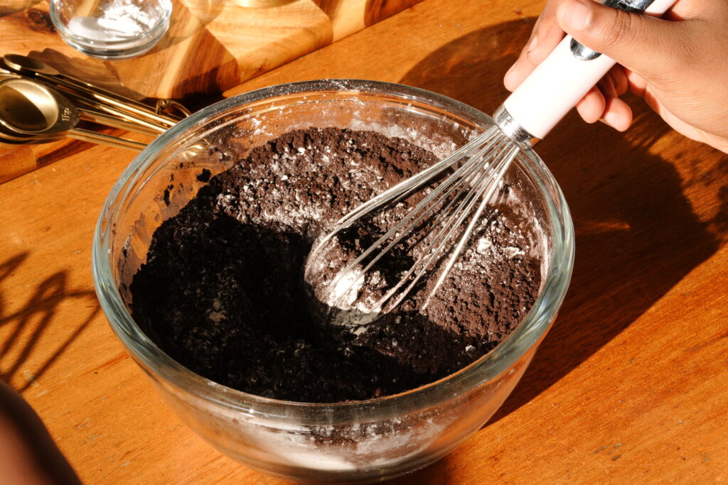 Mixing black cocoa powder and flour in a glass bowl using a whisk under warm lighting.