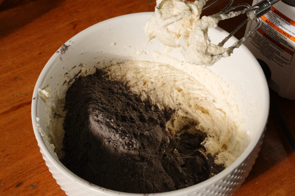Dry black cocoa mixture added to a creamy butter base in a mixing bowl with a hand mixer.