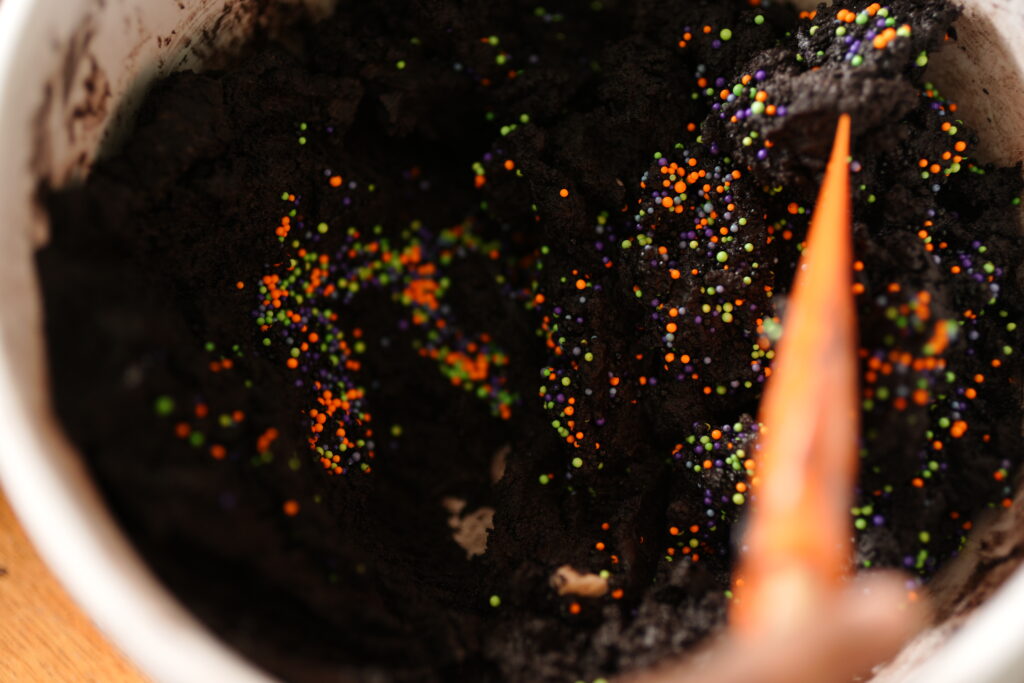 Hand mixing Halloween sprinkles into dark cocoa dough with an orange spatula.