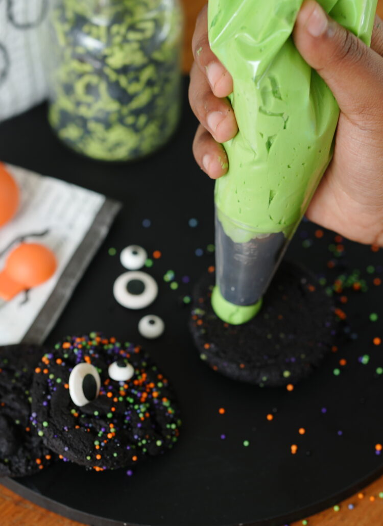 A child’s hand piping bright green frosting onto a black cocoa cookie surrounded by Halloween sprinkles and candy eyes.