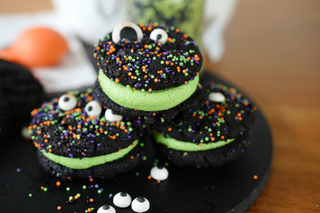 Stacked black cocoa cookie sandwiches filled with green frosting and topped with candy eyeballs and colorful Halloween sprinkles.