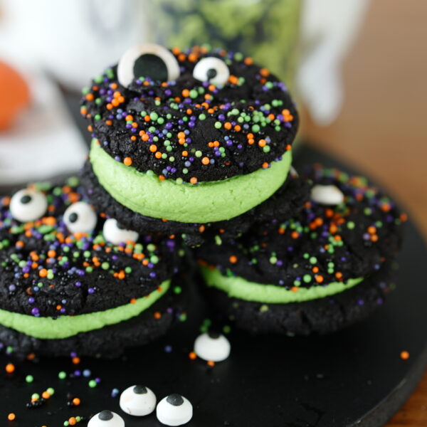 Stacked black cocoa cookie sandwiches filled with green frosting and topped with candy eyeballs and colorful Halloween sprinkles.