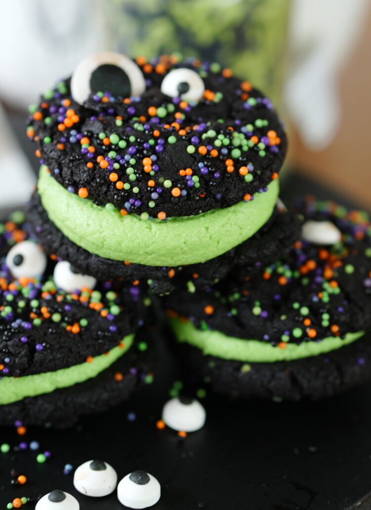 Stacked black cocoa cookie sandwiches filled with green frosting and topped with candy eyeballs and colorful Halloween sprinkles.