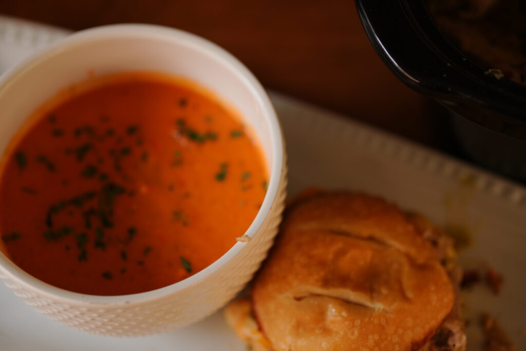 A bowl of creamy crock pot tomato soup garnished with herbs served alongside a jerk pork grilled cheese sandwich on a white plate.