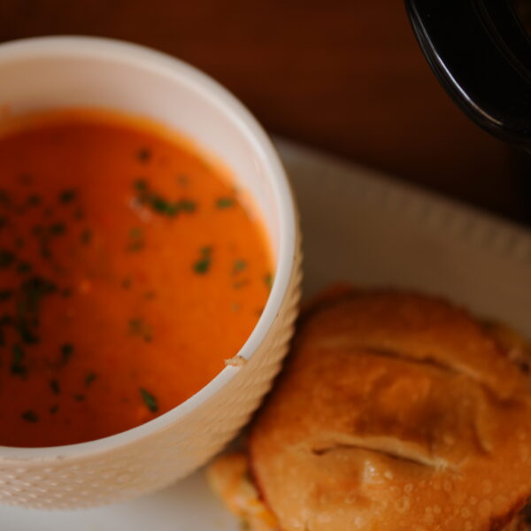 A bowl of creamy crock pot tomato soup garnished with herbs served alongside a jerk pork grilled cheese sandwich on a white plate.