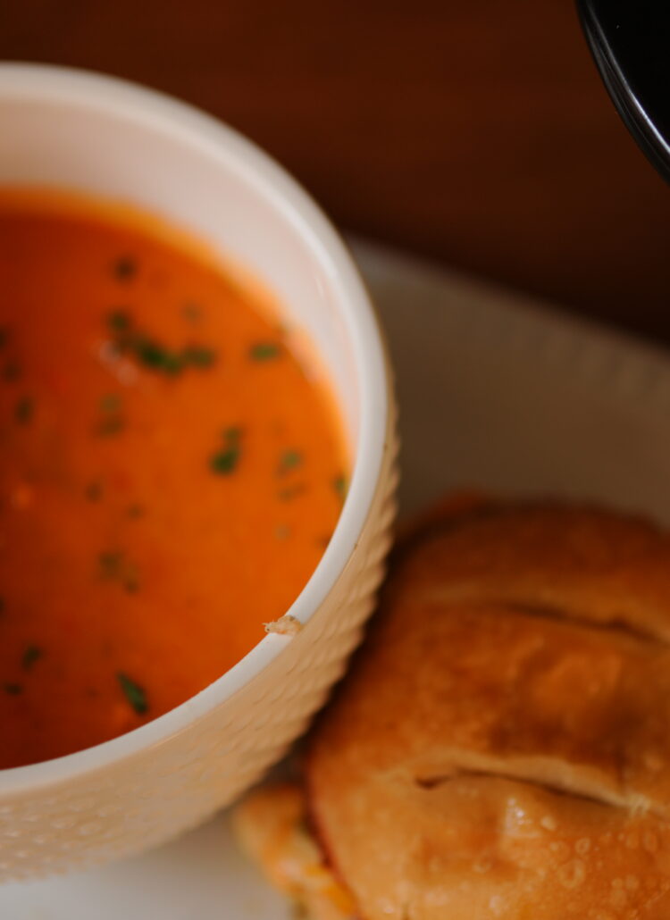 A bowl of creamy crock pot tomato soup garnished with herbs served alongside a jerk pork grilled cheese sandwich on a white plate.