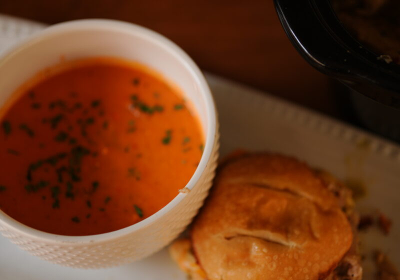 A bowl of creamy crock pot tomato soup garnished with herbs served alongside a jerk pork grilled cheese sandwich on a white plate.