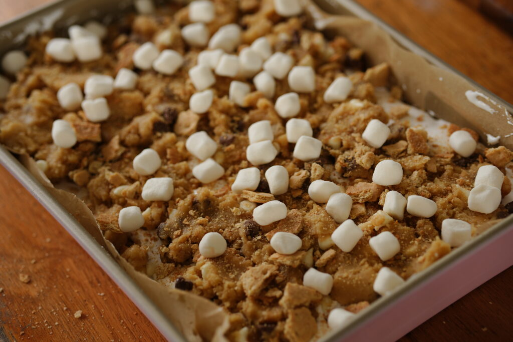 Baking pan topped with cookie dough pieces, mini marshmallows, chocolate chunks, and graham bits before baking.