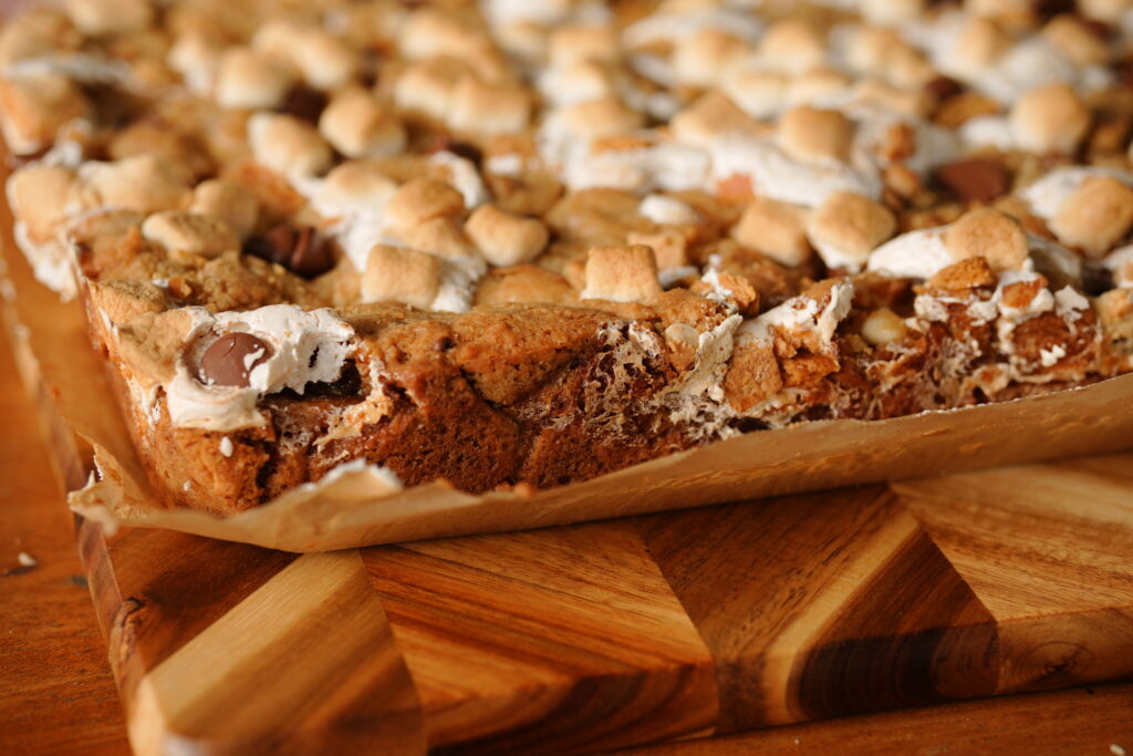 Close-up of homemade s’mores cookie bars with toasted marshmallows, graham cracker chunks, and melted chocolate chips on a wooden board.