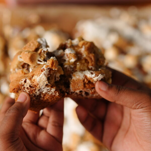 Hands holding a gooey s’mores cookie bar filled with marshmallows, melted chocolate, and graham crackers.