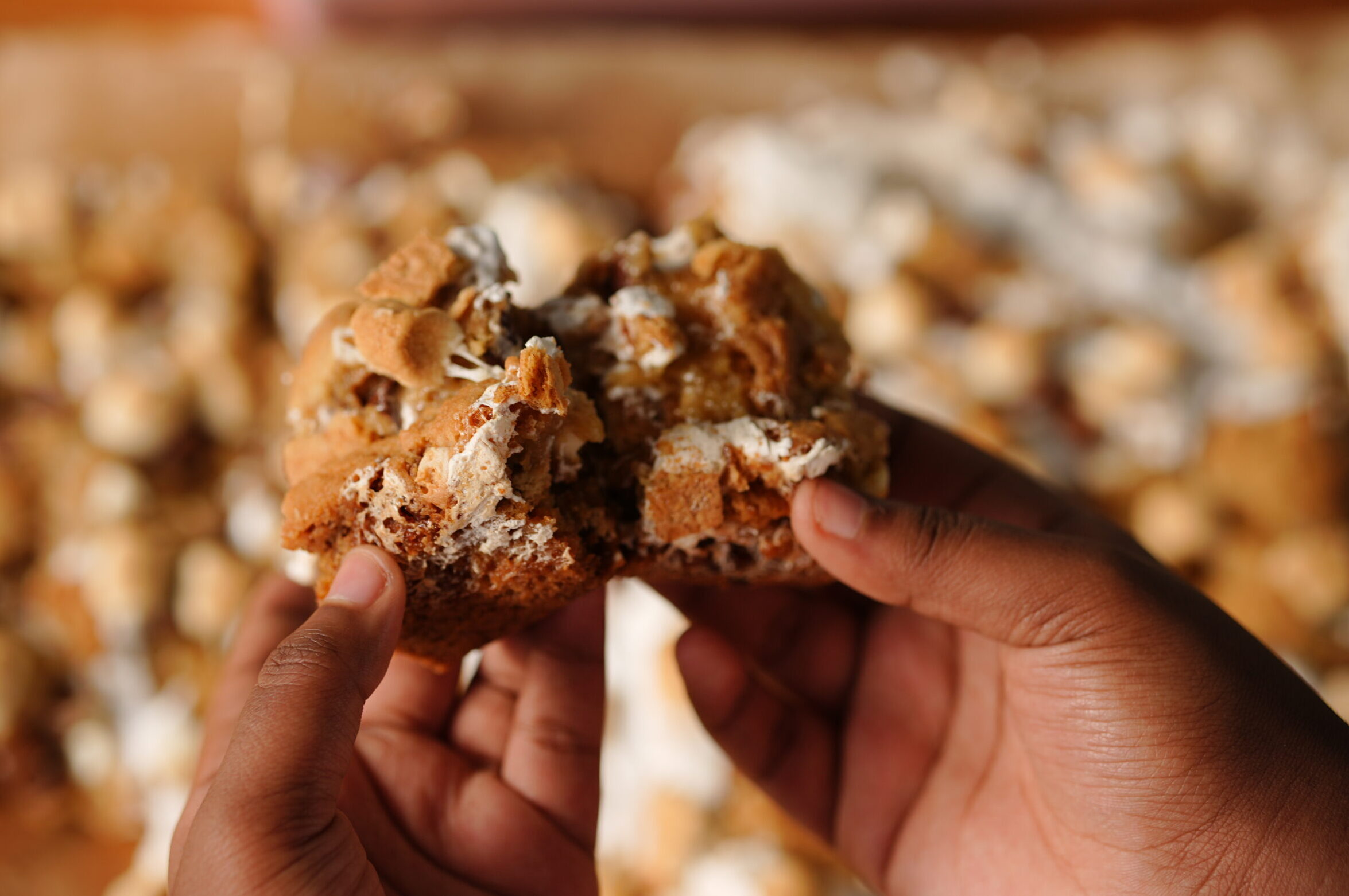 Hands holding a gooey s’mores cookie bar filled with marshmallows, melted chocolate, and graham crackers.