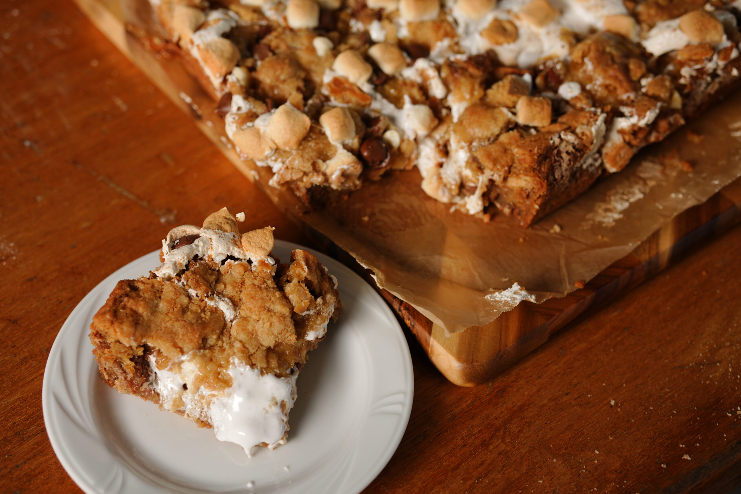 Close-up of a gooey homemade s’mores cookie bar with marshmallow, chocolate chips, and graham cracker crust, served on a white plate with the rest of the dessert in the background.