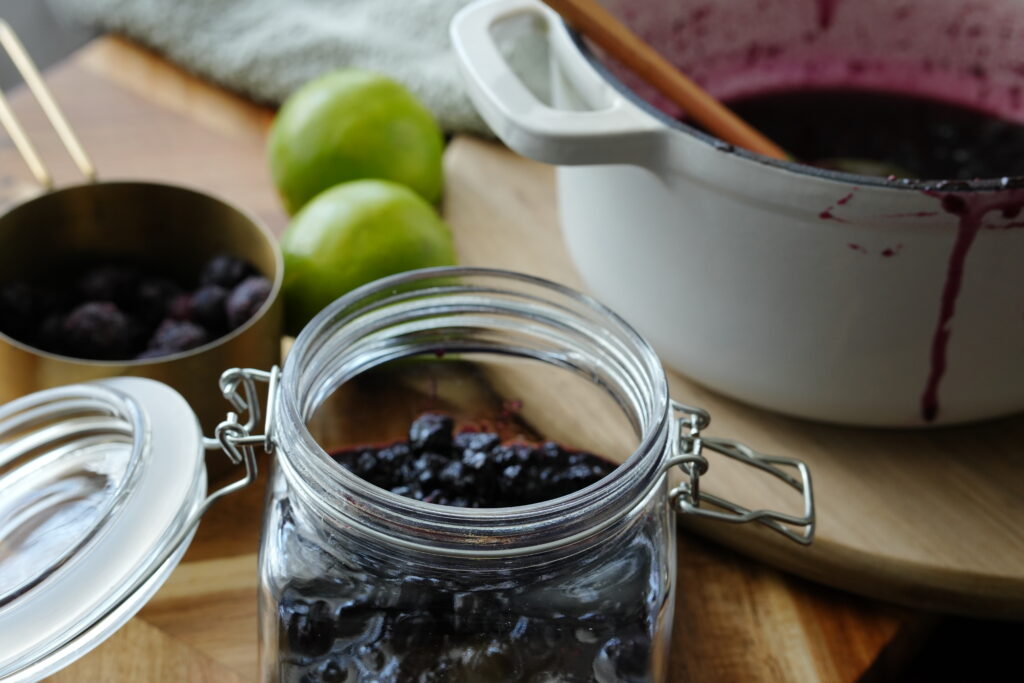 Glass jar of homemade blueberry jam on a wooden board with limes, a pot of cooked jam, and frozen blueberries in the background.