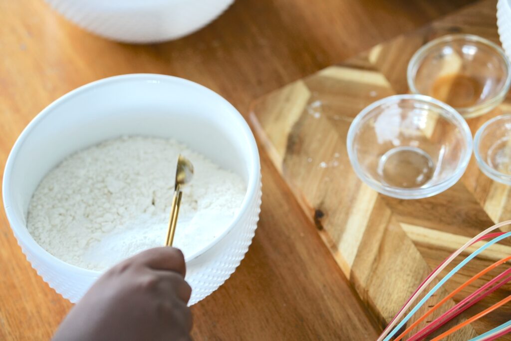 A hand holding a spoon over a bowl of flour, preparing the dry ingredients for white chocolate chip blueberry pie cookies.