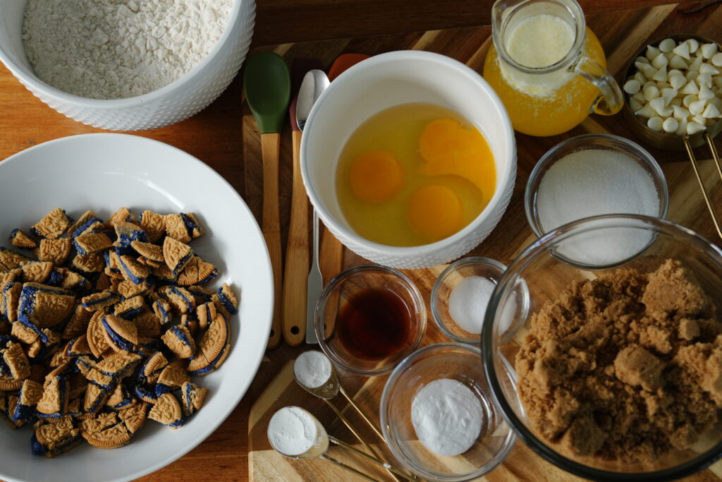 Overhead view of ingredients for white chocolate chip blueberry pie cookies, including eggs, Oreos, flour, and sugars.