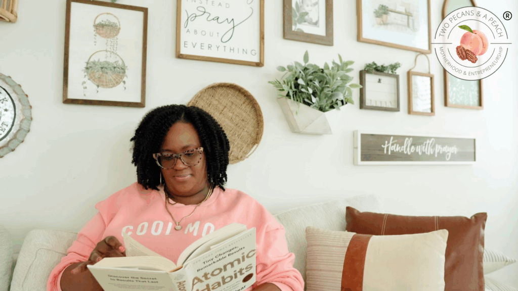 Black woman mompreneur in a pink sweatshirt reading Atomic Habits on a cozy living room couch embracing the benefits of Realistic Morning Routines for Entrepreneur Moms