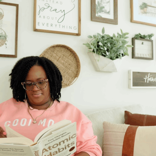 Black woman mompreneur in a pink sweatshirt reading Atomic Habits on a cozy living room couch