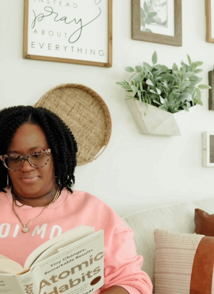 Black woman mompreneur in a pink sweatshirt reading Atomic Habits on a cozy living room couch