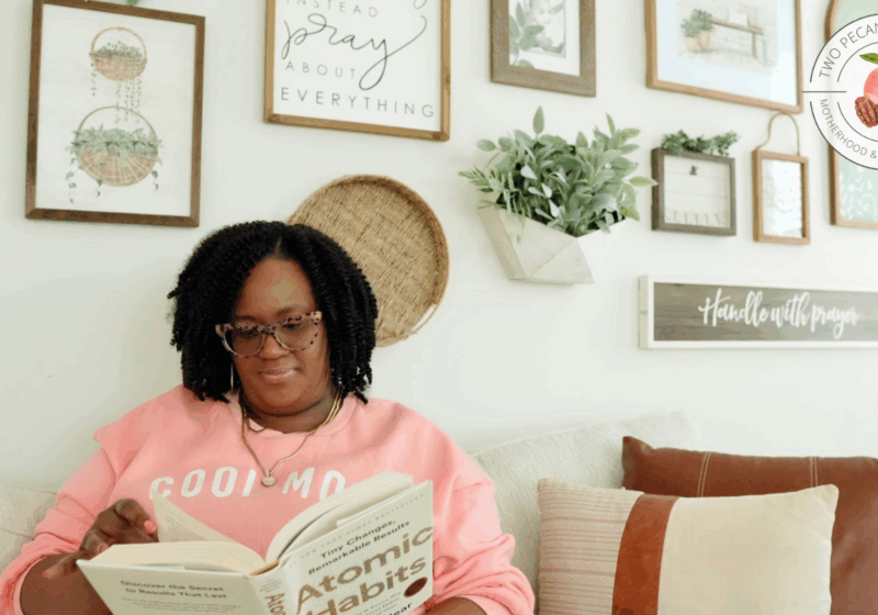 Black woman mompreneur in a pink sweatshirt reading Atomic Habits on a cozy living room couch
