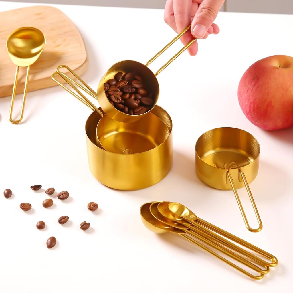 Set of gold stainless steel measuring cups and spoons with coffee beans, displayed on a white countertop with an apple and cutting board in the background.
