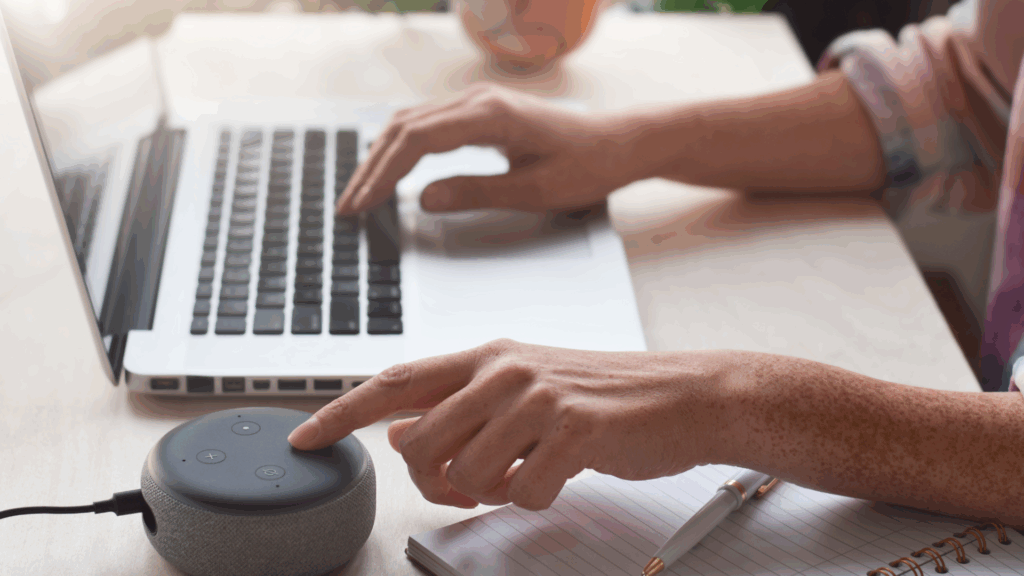 Hand activating a smart speaker next to a laptop and notebook, showing a streamlined tech-assisted morning setup.