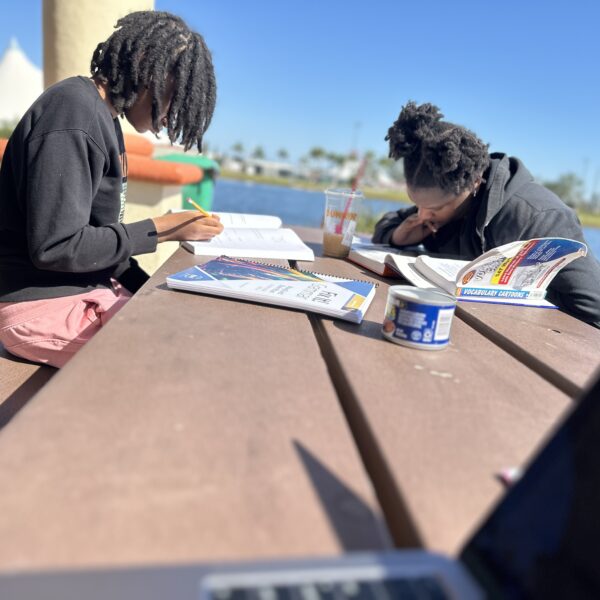 Two teenagers studying at a picnic table by the lake with homeschool books and iced coffee.