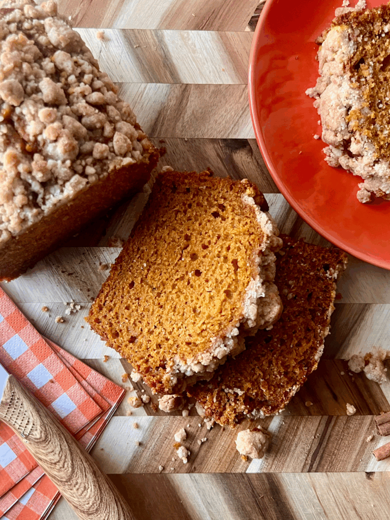 Sliced pumpkin bread with a golden pumpkin spice streusel topping, served on a wood cutting board with cinnamon sticks, a checkered napkin, and a white knife for a cozy fall aesthetic.