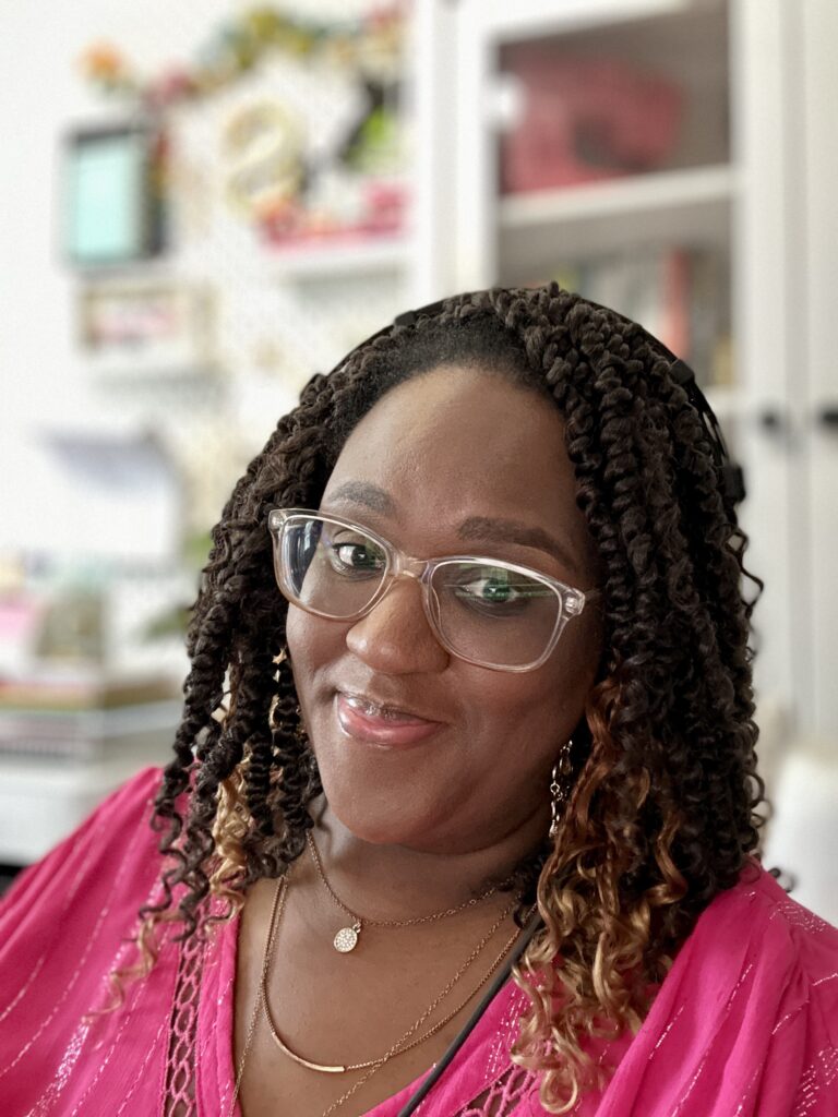 Shani Fay Allman smiling while seated at her desk, wearing a pink blouse and clear-framed glasses, preparing her podcast batching schedule