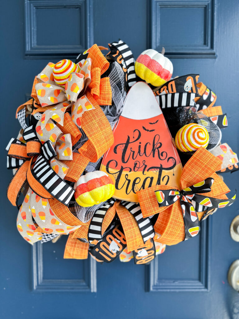 Halloween wreath on a blue door featuring candy corn-themed ribbon, plush candy decor, striped bows, and a Trick or Treat candy corn sign in the center