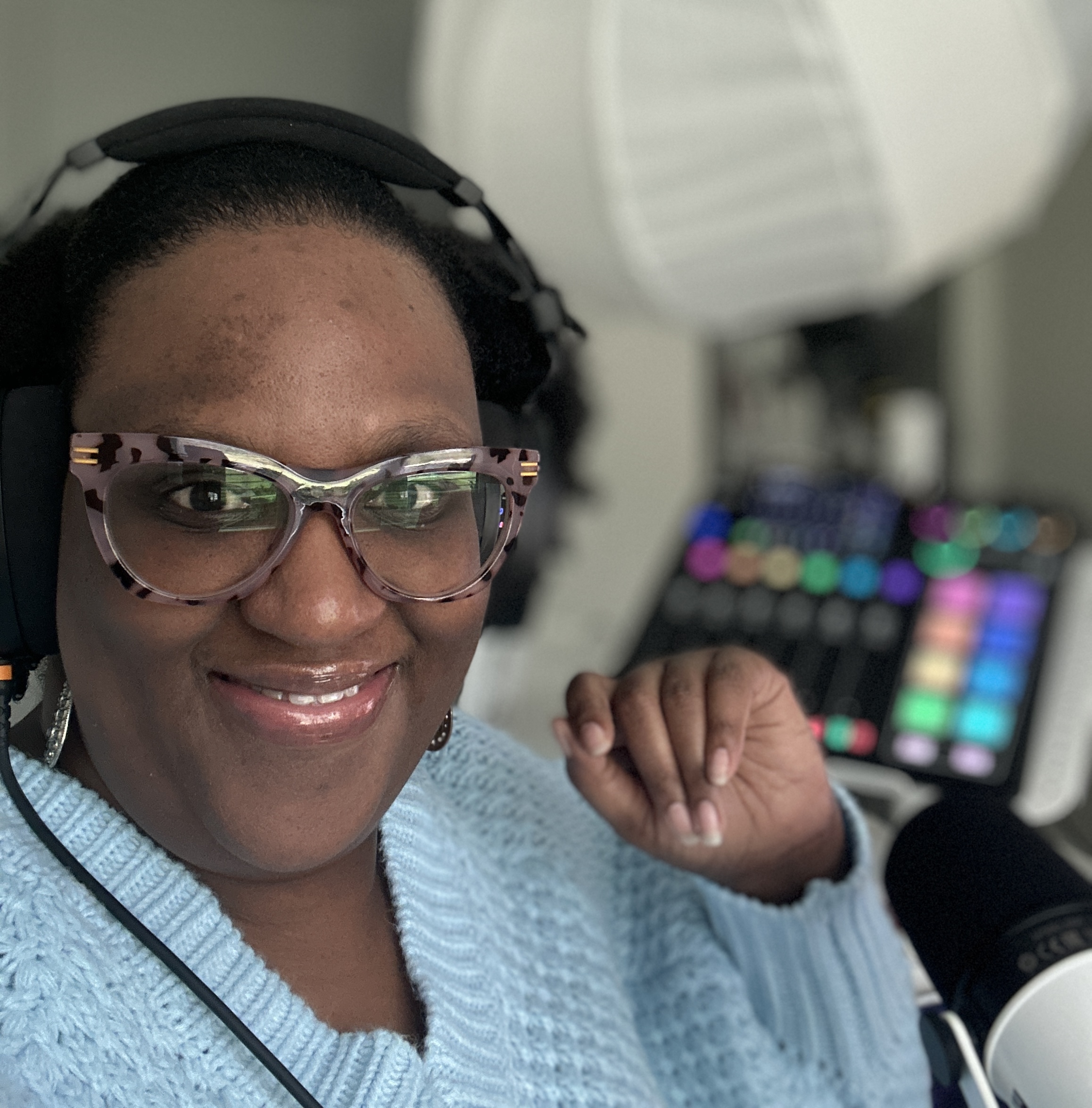 Shani Fay Allman recording a podcast in her home studio wearing headphones and glasses, with a colorful audio mixer and microphone visible.