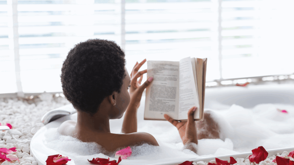 Black woman practicing self-care reading a book in a bubble bath with rose petals