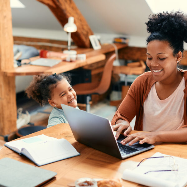 Mother smiling as she types on a laptop while her daughter stands beside her, laughing and enjoying the moment in a warm, lived-in workspace.
