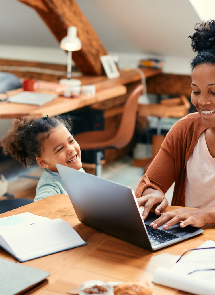 Mother smiling as she types on a laptop while her daughter stands beside her, laughing and enjoying the moment in a warm, lived-in workspace.
