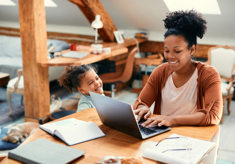 Mother smiling as she types on a laptop while her daughter stands beside her, laughing and enjoying the moment in a warm, lived-in workspace.