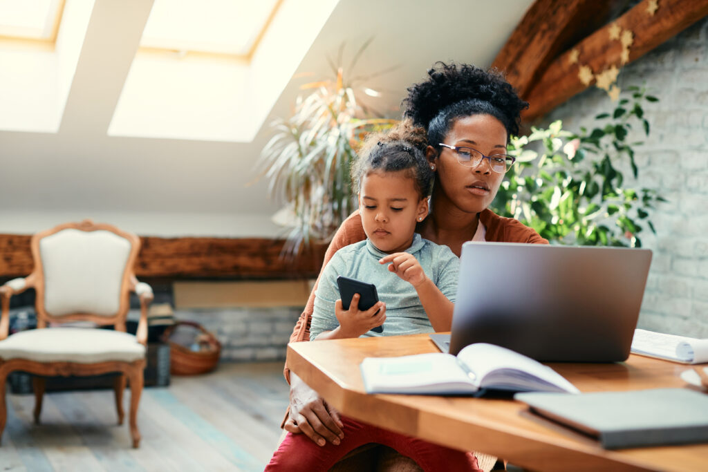 Black mother working from home on a laptop while her daughter sits in her lap holding a phone, both focused and engaged in a cozy, sunlit home office.