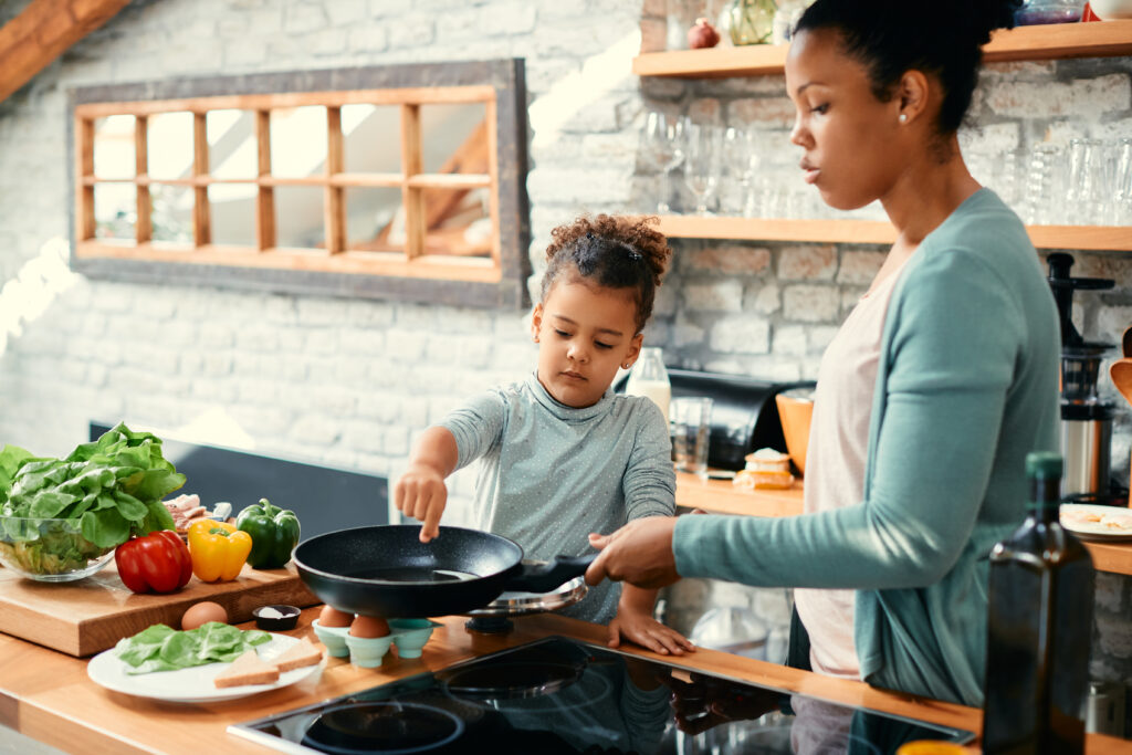 Young girl reaching into a skillet as her mother guides her at the stovetop; fresh produce and sandwich ingredients are spread out on the kitchen counter.