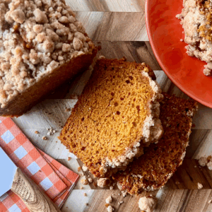 Sliced pumpkin bread with a golden pumpkin spice streusel topping, served on a wood cutting board with cinnamon sticks, a checkered napkin, and a white knife for a cozy fall aesthetic.