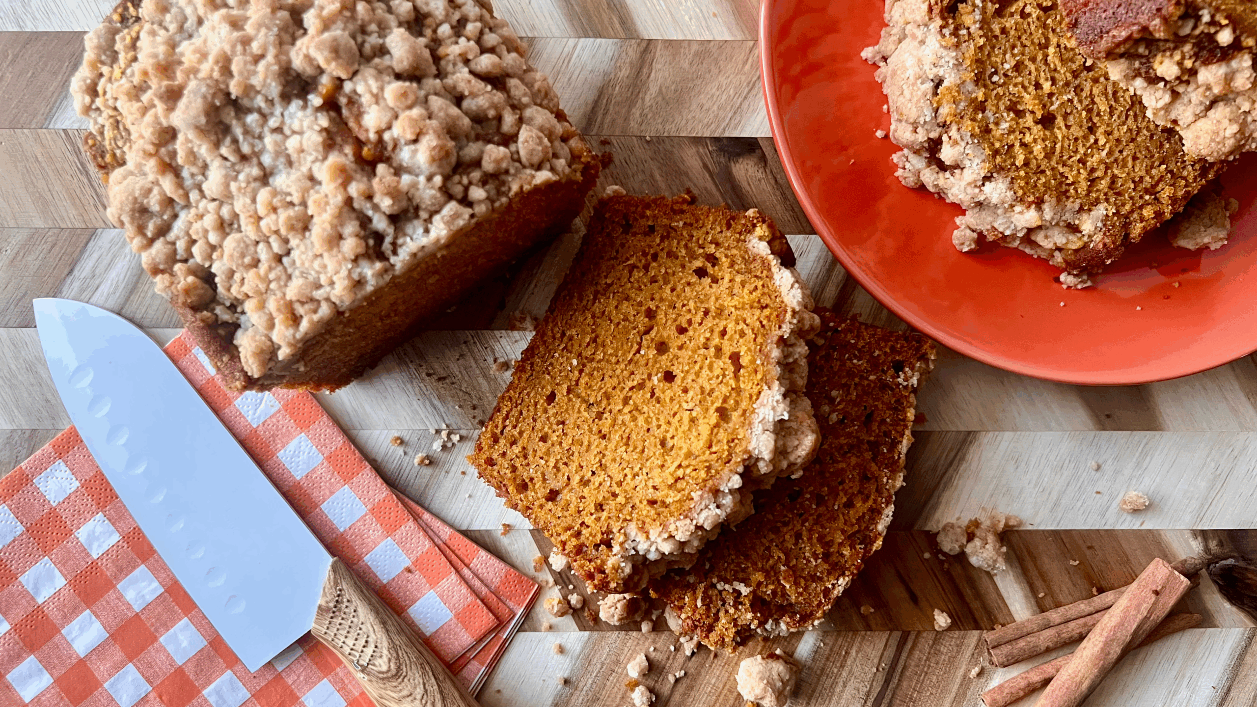 Sliced pumpkin bread with a golden pumpkin spice streusel topping, served on a wood cutting board with cinnamon sticks, a checkered napkin, and a white knife for a cozy fall aesthetic.