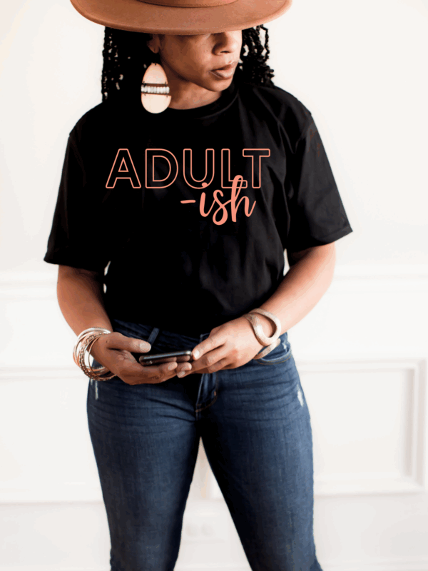 Woman standing in a black “Adult-ish” t-shirt, holding a phone, styled with jeans.
