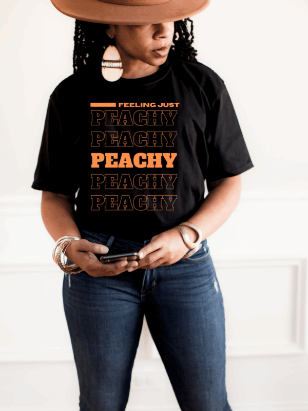 Woman in black “Feeling Just Peachy” t-shirt styled with jeans and wide-brim hat, looking down while holding phone