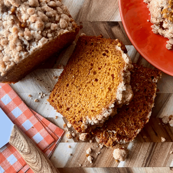 Sliced pumpkin bread with a golden pumpkin spice streusel topping, served on a wood cutting board with cinnamon sticks, a checkered