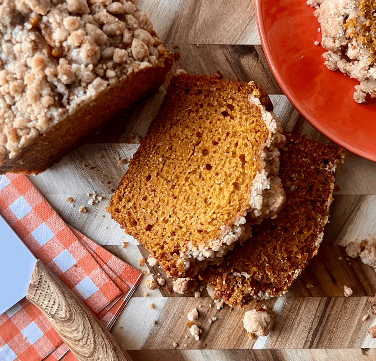 Sliced pumpkin bread with a golden pumpkin spice streusel topping, served on a wood cutting board with cinnamon sticks, a checkered