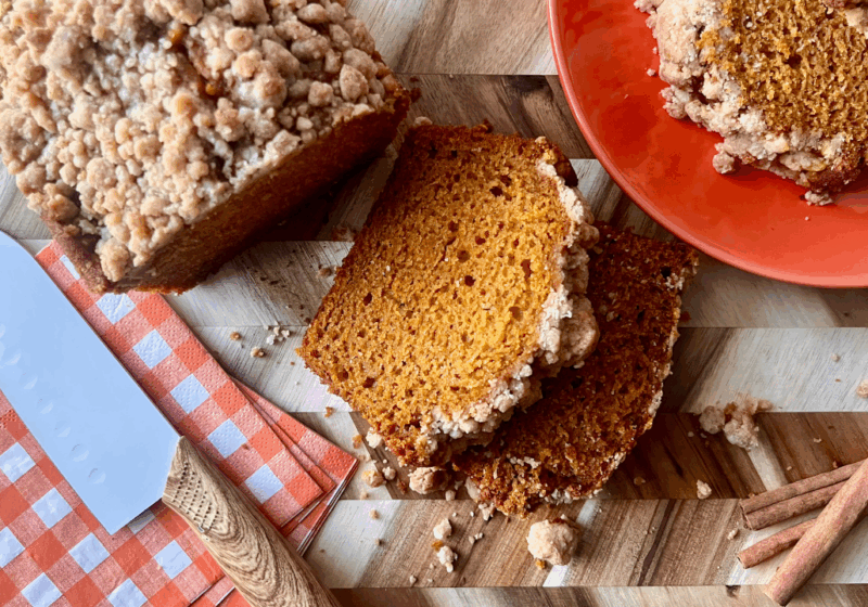 Sliced pumpkin bread with a golden pumpkin spice streusel topping, served on a wood cutting board with cinnamon sticks, a checkered