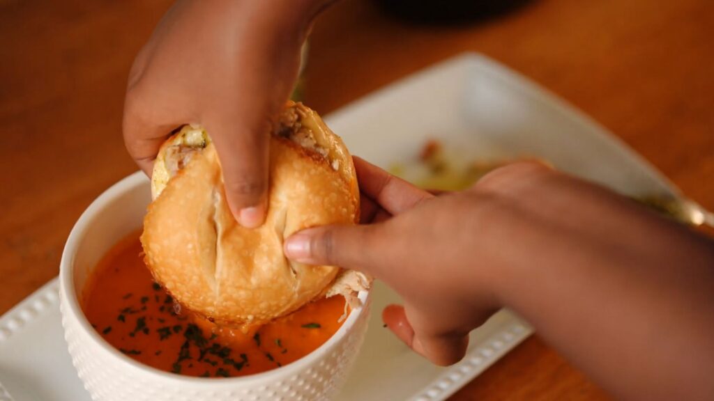 Hands dipping a jerk pork grilled cheese sandwich into creamy crock pot tomato soup topped with parsley.