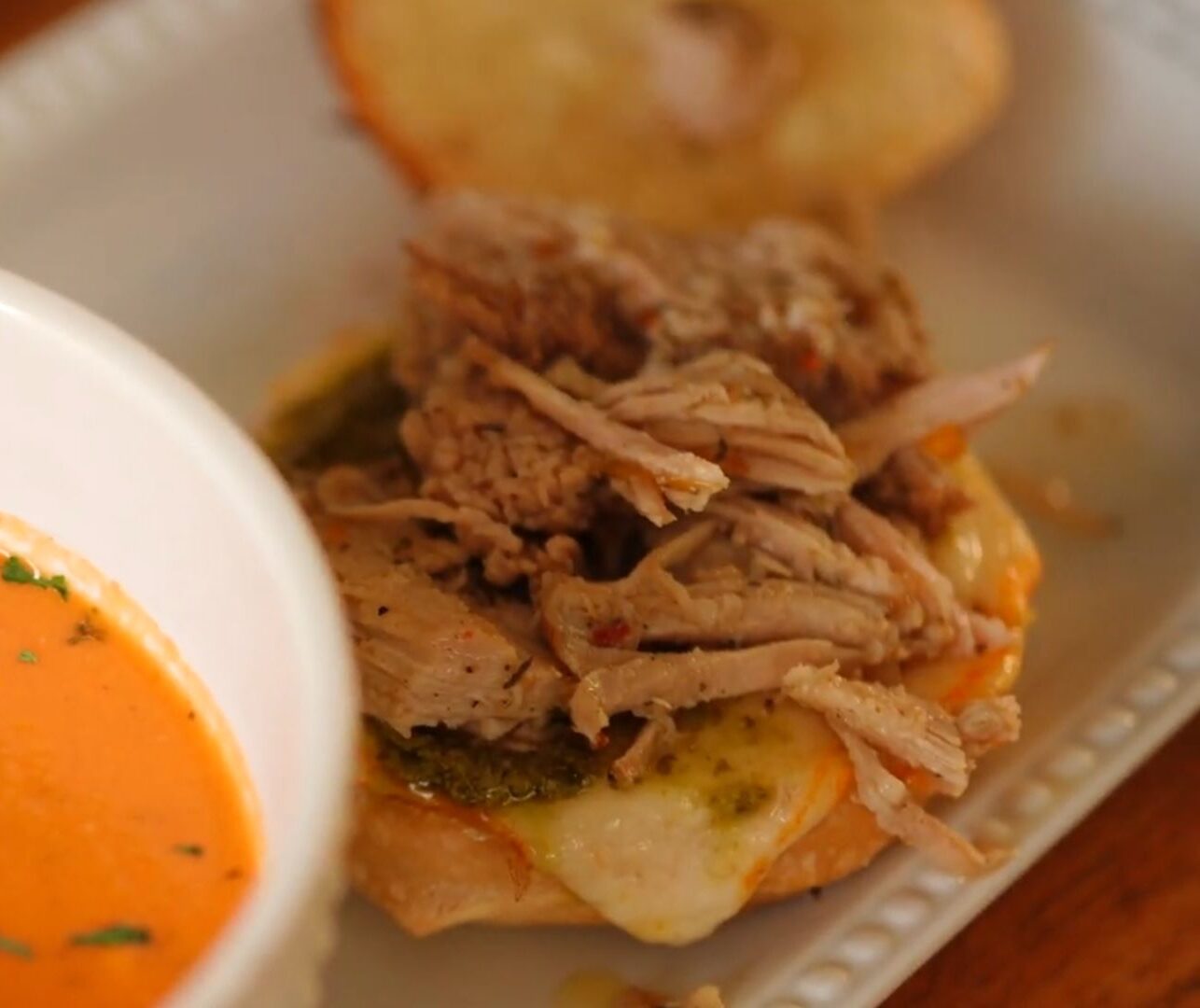 White bowl of creamy crock pot tomato soup served alongside a jerk pork grilled cheese sandwich.