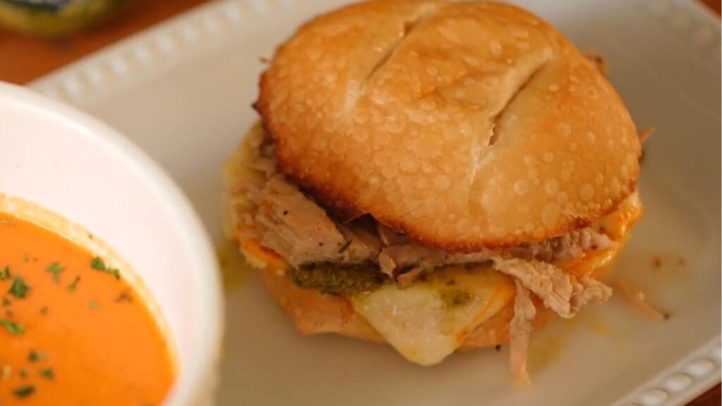 White bowl of creamy crock pot tomato soup served alongside a jerk pork grilled cheese sandwich.