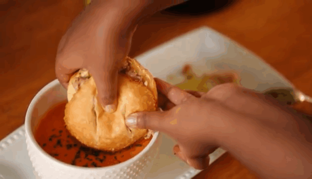 Hands dipping a jerk pork grilled cheese sandwich into a bowl of creamy crock pot tomato soup with fresh herbs.