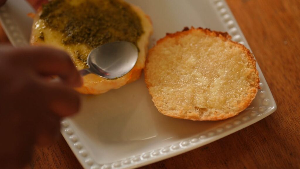 Close-up of bread topped with melted cheese and pesto, ready to be layered with jerk pork.