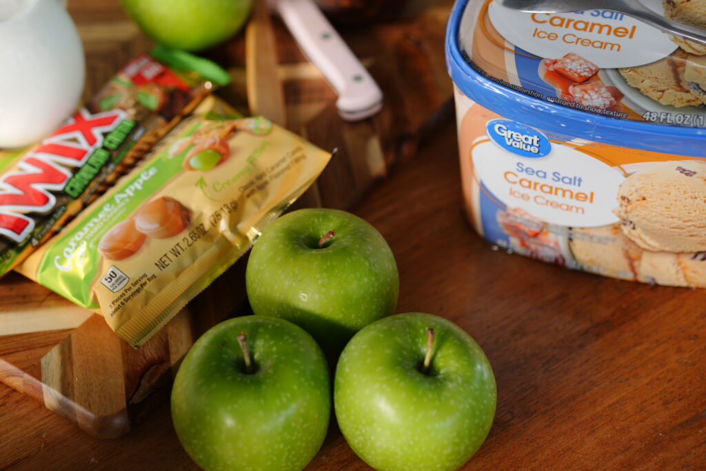 A close-up of Granny Smith apples, caramel apple Twix, and Great Value Sea Salt Caramel Ice Cream arranged on a wooden table for a homemade caramel apple milkshake recipe.